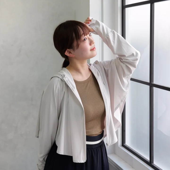 窓の外を眺める若い女性のポートレート。白いパーカーと茶色のトップスを着ており、日差しを避けるように手を額にかざしている。