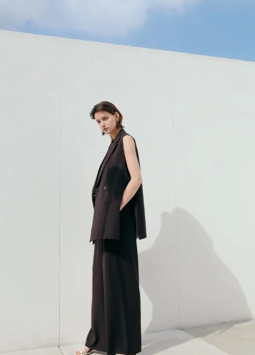 ダークカラーのノースリーブジャケットとワイドパンツを着用した女性の全身ショット。ミニマルで洗練されたファッションスタイル。