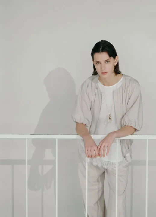 淡いグレーのジャケットとパンツを着用し、白いレールに手を置いて立つ女性。ミニマルな雰囲気のファッション写真。