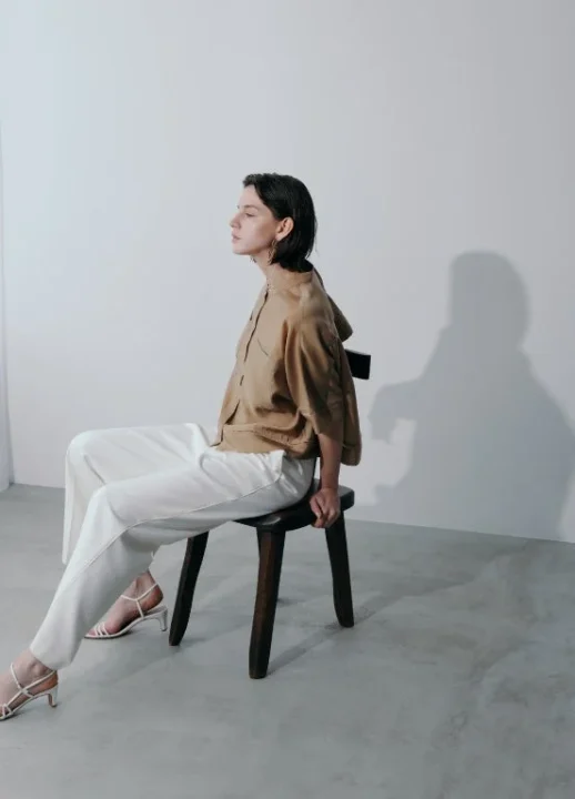 ベージュのトップスと白いパンツを着用した女性が木製スツールに座っている。ミニマルな雰囲気。