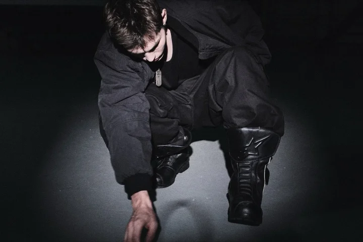 暗い背景の中、黒い服と特徴的なブーツを着用した人物がしゃがんでいる俯瞰ショット。クールでミステリアスな雰囲気が漂う。