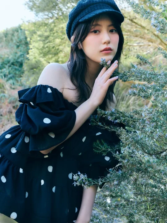 黒い水玉のオフショルダーワンピースと帽子を身につけた女性が、屋外の緑豊かな植物に囲まれ、花にそっと触れているポートレート