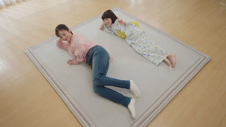 明るいリビングのラグの上で、女性と浴衣姿の女の子が仲良く横になっています。二人は笑顔でくつろいでおり、穏やかな家族のひとときが感じられます。