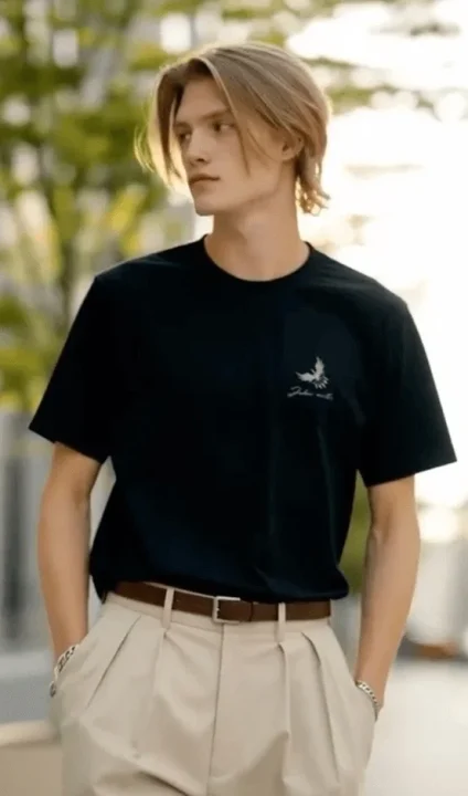 金髪の長髪の男性が黒いTシャツとベージュのプリーツパンツを着用し、屋外でカジュアルなポーズをとっている。