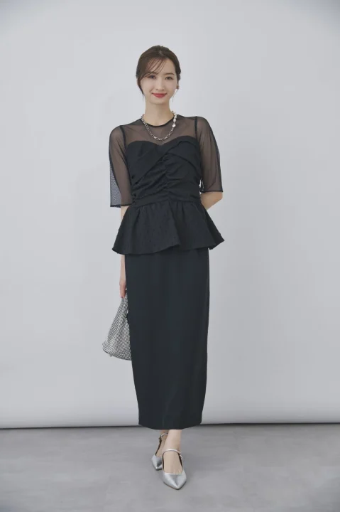 黒いシースルートップスとペプラムデザインが特徴のロングドレスを着用した女性が、スタジオで微笑んでいます。