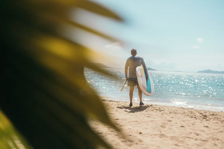 パドルボードとパドルを持った男性がビーチに立ち、きらめく海を眺めている