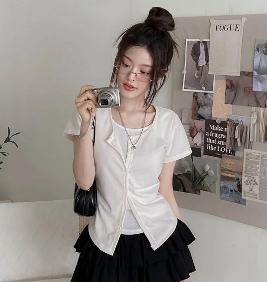 お団子ヘアと眼鏡の女性が、白いトップスと黒いミニスカートを着用し、銀色のデジタルカメラを構えている。