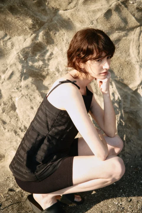 ショートヘアの女性が黒い服を着て、岩肌を背景にしゃがみ込んでいるポートレート。