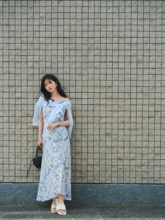 青い柄のロングワンピースを着た女性が、グレーのタイル壁にもたれかかって立っています。肩に羽織った水色のカーディガンと白いサンダルが、軽やかでエレガントな夏のファッションを演出しています。