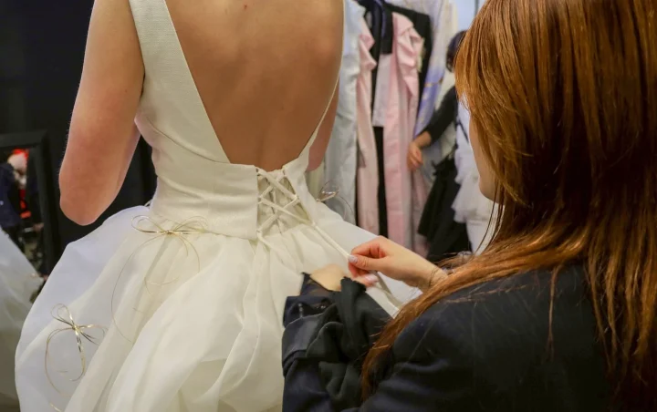 白いウェディングドレスを着た花嫁の背中を、別の人が編み上げ式の紐で調整している場面です。結婚式の準備中のようです。