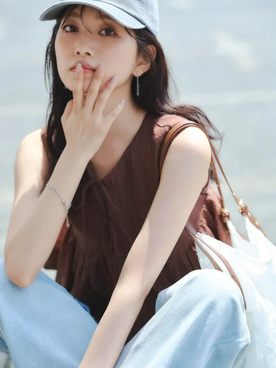 キャップをかぶり、茶色のノースリーブと薄いブルージーンズを着用した若い女性が屋外でポーズをとっています。片手を口元に添え、カメラを見つめる表情が印象的で、カジュアルながらも洗練された夏のファッションスタイルを表現しています。