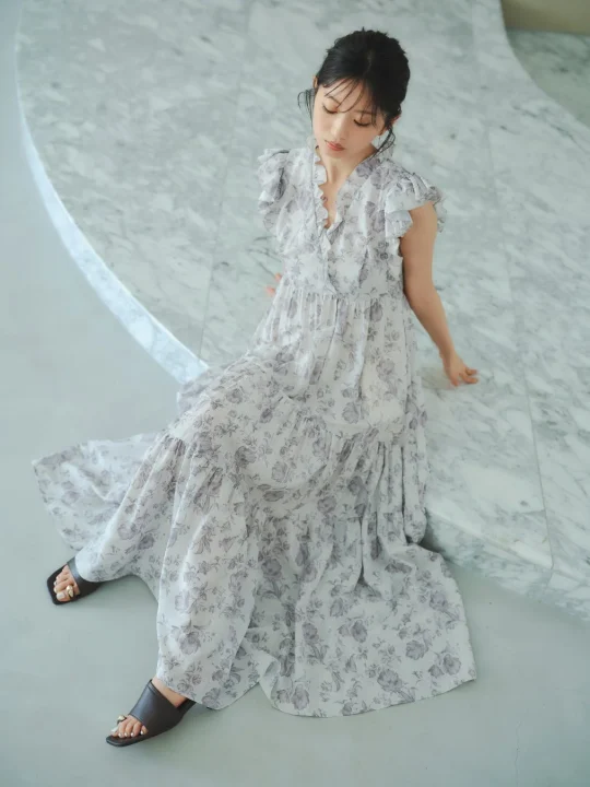 白い花柄のマキシ丈ワンピースを着た女性が大理石の床に座り、優しくうつむいています。フリルとティアードデザインが特徴のワンピースは、エレガントでフェミニンな雰囲気を醸し出しています。