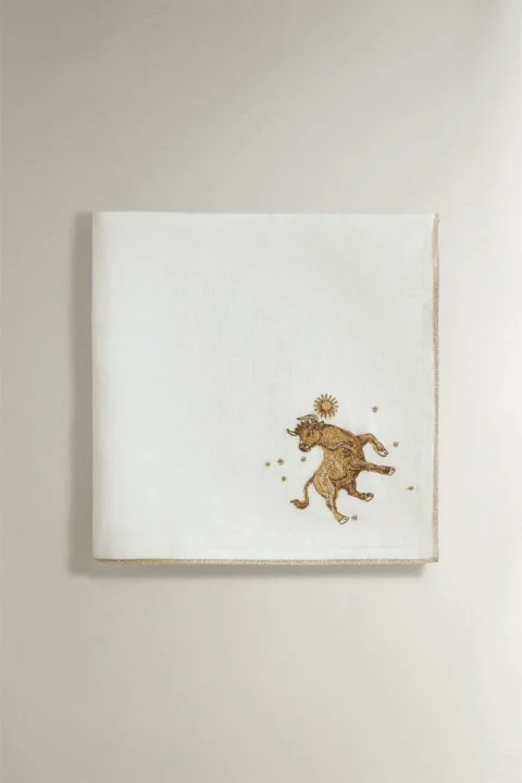 白いリネン生地に、雄牛と太陽、星をモチーフにした金色の刺繍が施された布製品です。縁には金色のトリムがあり、エレガントな印象を与えます。