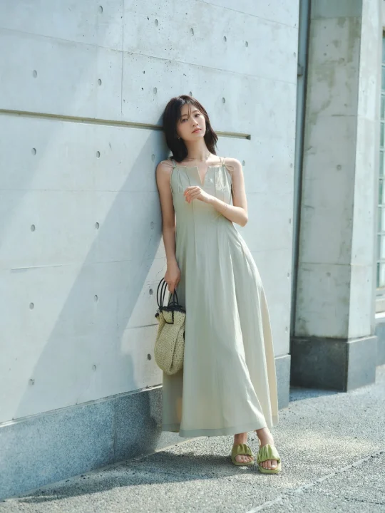 コンクリートの壁に寄りかかる若い女性の全身像です。淡いグリーンのロングワンピースに麦わらバッグとサンダルを合わせた、夏らしいリラックスしたカジュアルファッションを着こなしています。明るい日差しの中で自然体な雰囲気です。