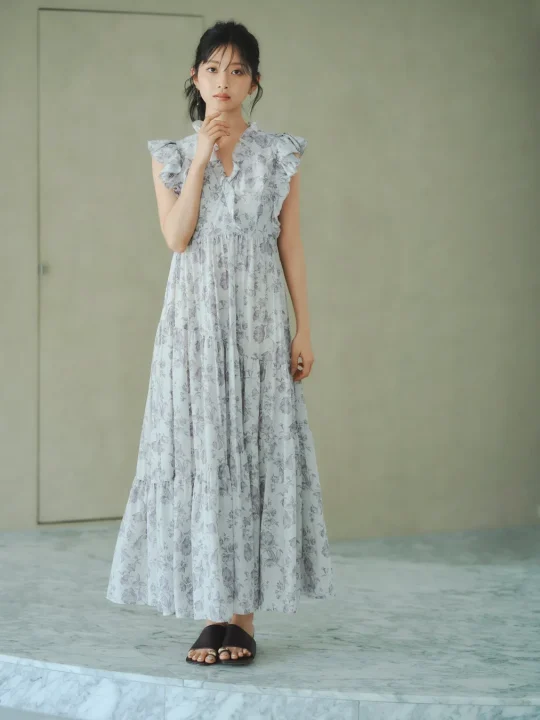 薄い花柄のフリル付きマキシワンピースを着た若い女性が、シンプルな背景の屋内で顎に手を当てて立っています。上品で涼しげな夏のファッションスタイルです。