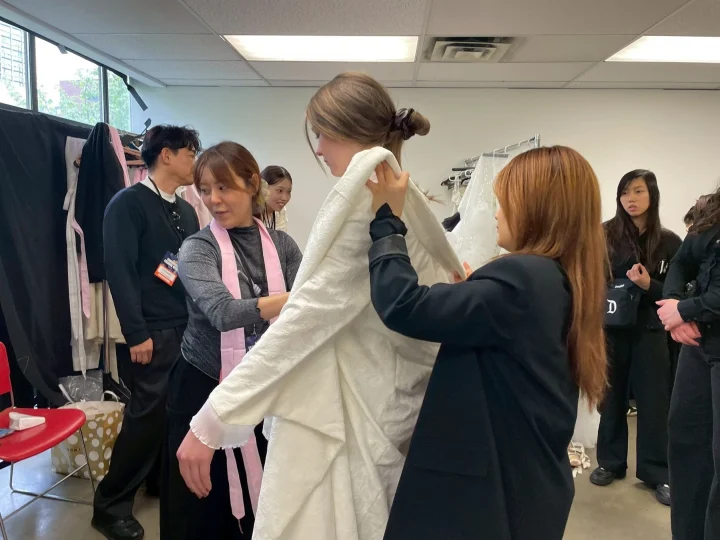 ファッションイベントのバックステージで、スタッフやボランティアがモデルの女性に白いドレスをフィッティングさせている様子です。他の関係者も周囲で見守り、イベントの準備が進められています。