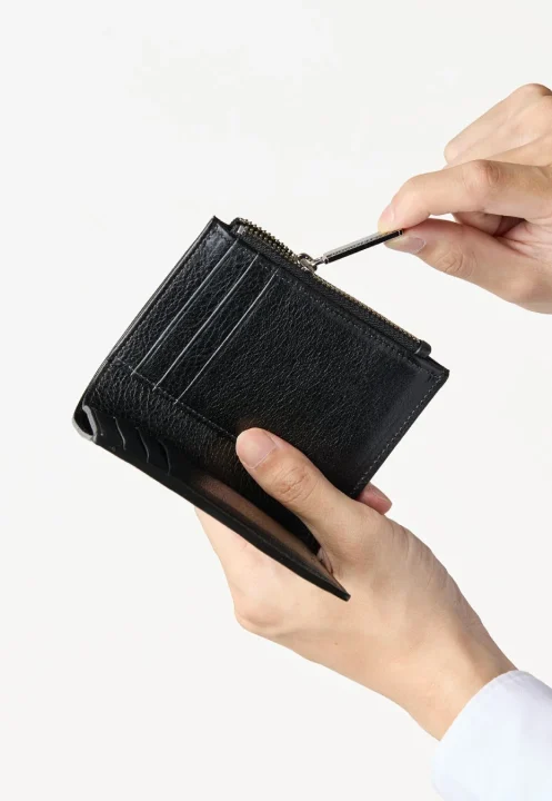 白い背景の前で、両手で黒い革製の財布を持ち、ジッパーを開けようとしている人物の様子を捉えた画像です