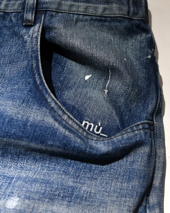 青いデニムジーンズのポケット部分のクローズアップ画像。ポケットの縁には「mū_」というロゴが刺繍されており、生地には白いペンキのような飛沫や軽いダメージ加工が見られます。