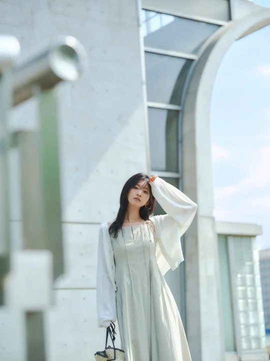 現代的な建築物を背景に、淡い色のワンピースと白いカーディガンを着用した若い女性がポーズをとっている夏のポートレートです。