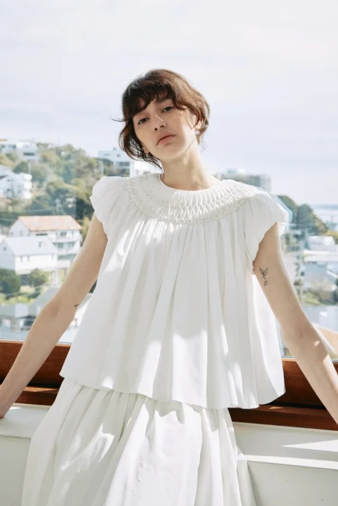 白いスモックブラウスとスカートを着用した女性が、明るい日差しの下、バルコニーでポーズをとっています。