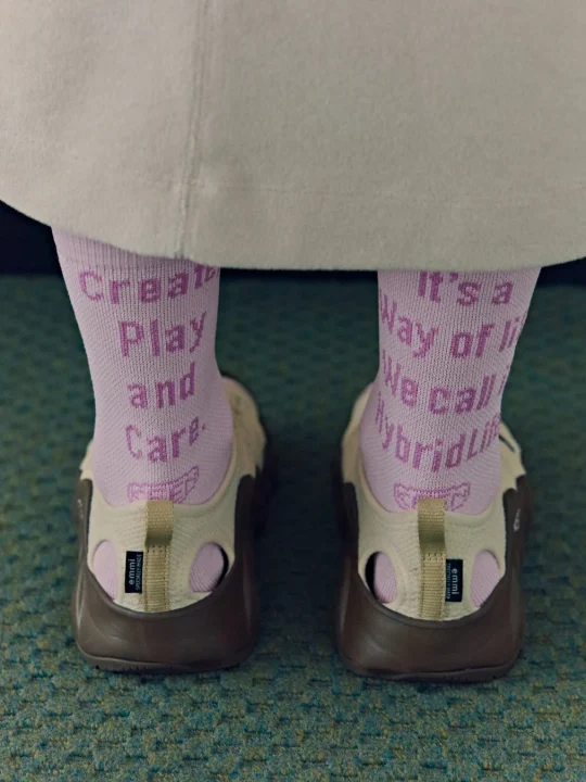 ピンクのソックスと茶色とベージュのemmiサンダルを履いた足元を捉えた写真。ソックスには「Create. Play. And Care. It's a Way of life. We call HybridLife.」の文字が見える。