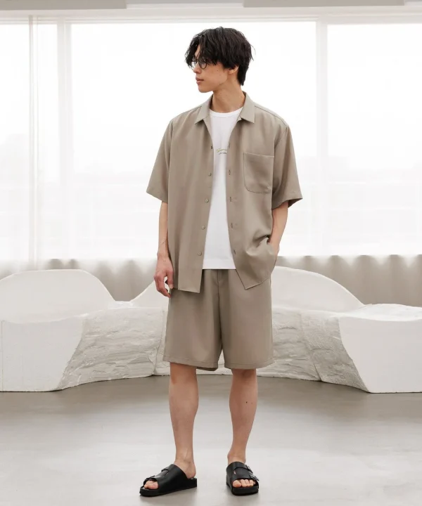 ベージュの半袖シャツとショーツのセットアップを着用した男性が、室内でポーズを取っている。リラックスした雰囲気の夏のカジュアルスタイル。