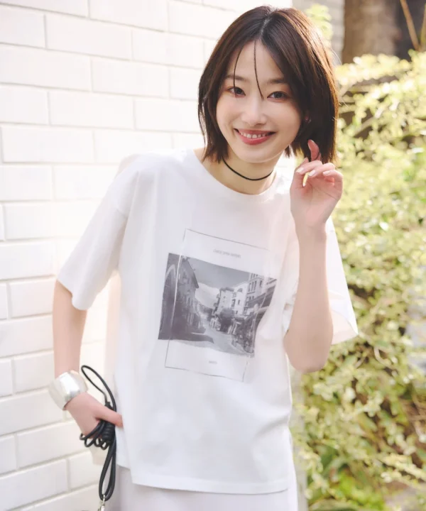 白Tシャツを着たショートヘアの若い女性が屋外で笑顔を見せています。Tシャツには建物のモノクロ写真と「CHASE OPEN WATERS」「PARIS」の文字がプリントされており、カジュアルで明るい雰囲気のポートレートです。