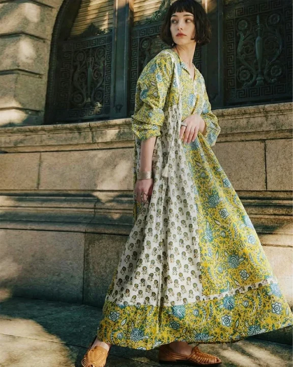黄色と白を基調としたエスニックな花柄のロングワンピースを着た女性が、歴史的な建物の前で立っています。