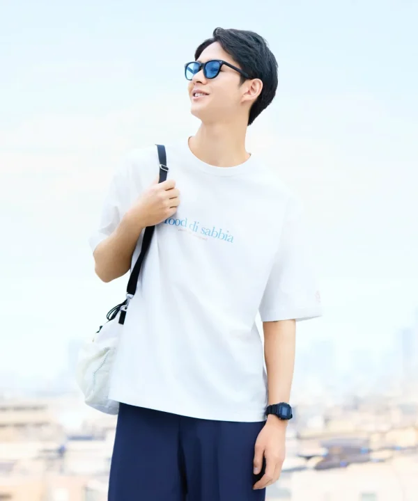 白いTシャツと青いサングラスを身につけた若い男性が、明るい屋外で爽やかに微笑んでいます。肩掛けバッグと腕時計を身につけ、リラックスした夏のカジュアルスタイルを見せています。