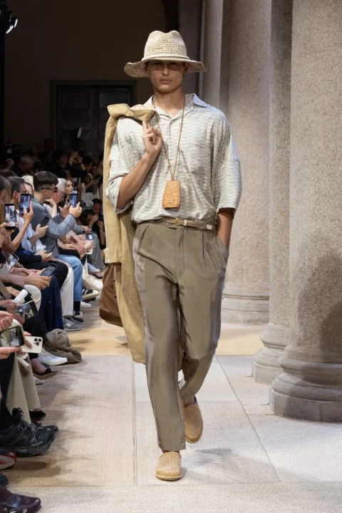 ファッションショーのランウェイを歩く男性モデル。ストローハット、柄シャツ、ベージュのパンツ、編み込みの靴を着用し、肩にジャケットを掛けています。