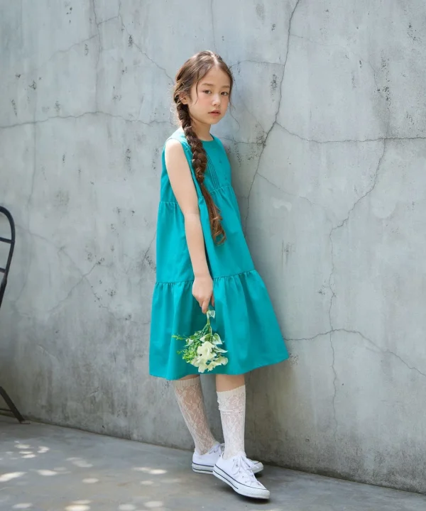 ターコイズブルーのワンピースを着た少女が、コンクリートの壁にもたれかかり、白い花束を持っている。編み込みヘアとレースのソックスが可愛らしい、屋外でのポートレート写真。