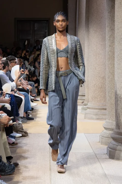 ランウェイを歩くモデルが写っています。彼女は光沢のある模様のジャケット、きらめくクロップトップ、そして淡いブルーのゆったりとしたパンツを着用しています。