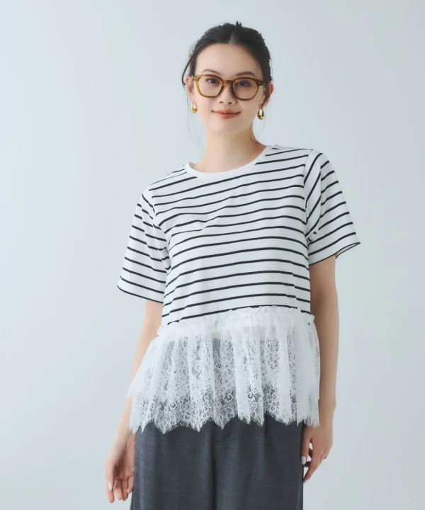 白い背景の前に立つ、髪をまとめた女性のポートレートです。黒と白のボーダーTシャツに白いレースの裾飾り、濃いグレーのパンツを着用し、茶色のメガネとゴールドのピアスを身につけています。穏やかな表情でカメラを見ています。