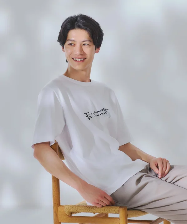 白い背景のスタジオで、木製の椅子に座って微笑む若い男性のポートレート。白のTシャツと淡い色のパンツを着用し、爽やかな印象を与えている。