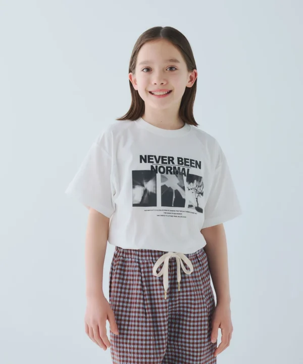 白いTシャツとチェック柄のパンツを着用した若い女の子が、白い背景の前で笑顔で立っています。Tシャツには「NEVER BEEN NORMAL」と書かれています。