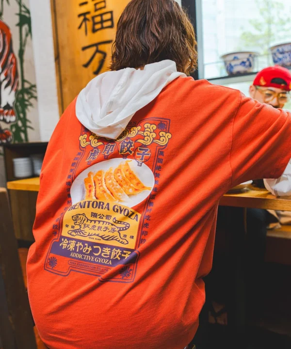 背中を向けた人物が、餃子のイラストと「BENITORA GYOZA」「冷凍やみつき餃子」などの文字がデザインされたオレンジ色のTシャツを着ている。店内のような場所で、奥には別の人物も見える。