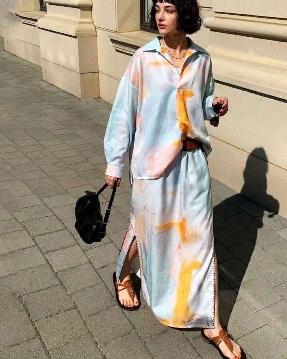 日差しの当たる街路を歩くショートヘアの女性。水彩画のような柄のツーピースを着て、サンダルと小さなバッグを合わせています
