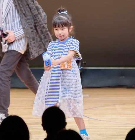 青と白のストライプのワンピースを着た幼い女の子が、青いぬいぐるみを持ち、笑顔でステージに立つ