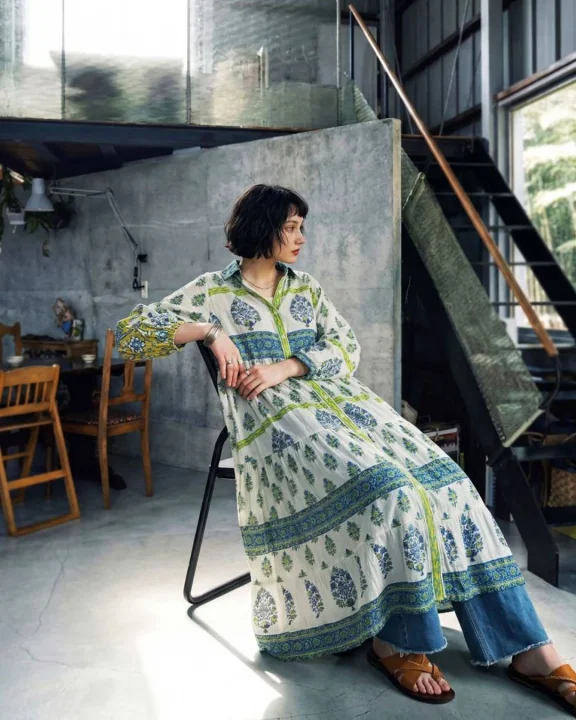 ショートヘアの女性が、柄物のロングワンピースにデニムを重ね着し、サンダルを履いて椅子に座っている