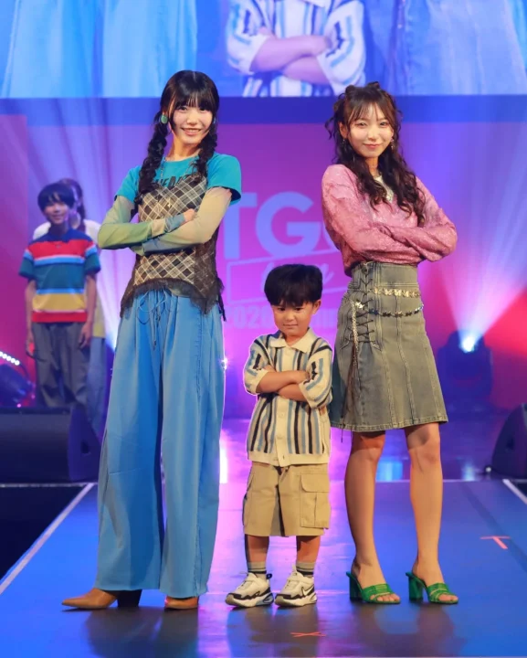 ファッションショーのステージで、2人の女性モデルと1人の子供が笑顔でポーズをとっています。
