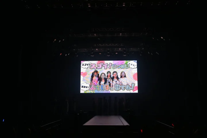 暗い会場のステージに設置された大型スクリーンに、5人の女の子たちが映し出されています。