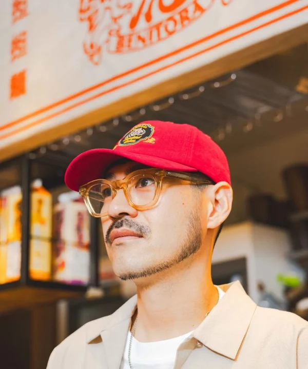 赤い帽子と眼鏡をかけた男性が、店内の看板を見上げている横顔のポートレート。口ひげと顎ひげが特徴的で、カジュアルな服装をしている。背景には「紅虎 NITORA」の文字が見える。