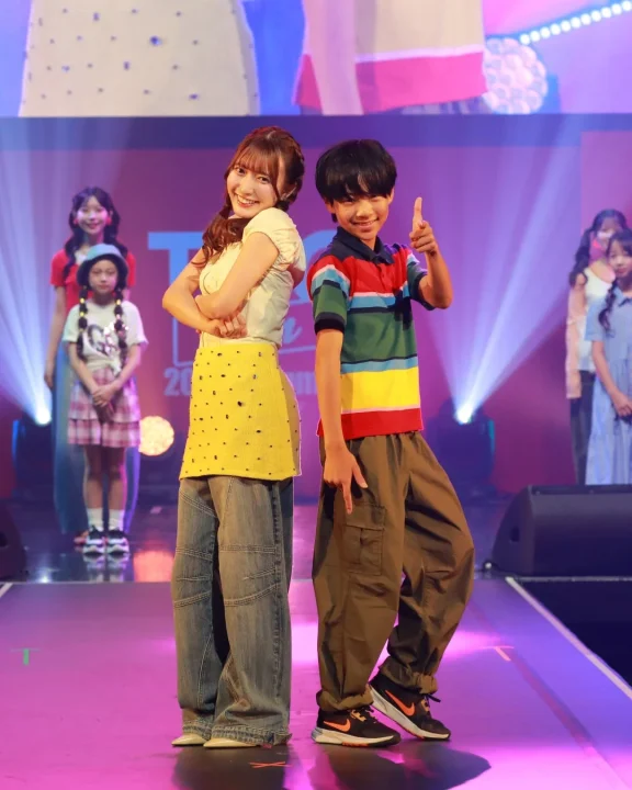 明るい照明のランウェイで、笑顔の若い女性と男の子がカジュアルな服装でポーズをとっています。