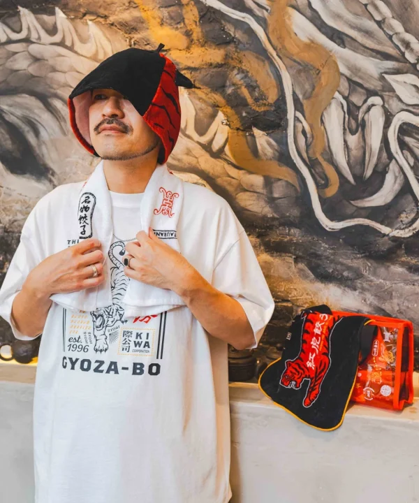 独特な帽子をかぶり、名物餃子と虎のデザインが施されたTシャツとタオルを身につけた男性が写っています。背景には抽象的な壁画があり、右には紅虎餃子房のロゴ入り虎柄グッズが見られます。