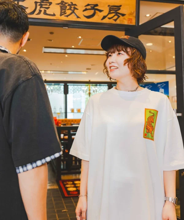 虎餃子房の入口付近で、虎柄のTシャツとキャップを身につけた女性が笑顔で男性と会話している様子です。明るい店内で、楽しそうな雰囲気が伝わってきます。