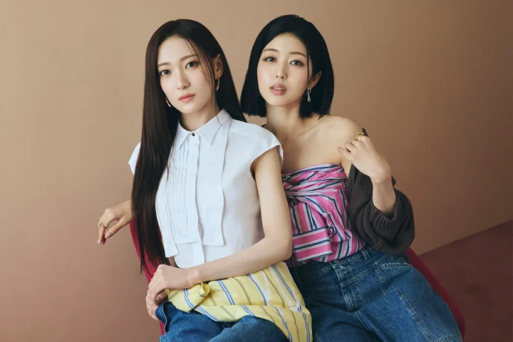 茶色の背景の前で、白いシャツとストライプのトップスを着た2人の女性が座ってポーズをとっている。モダンでおしゃれな雰囲気のポートレート写真。