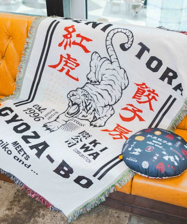 オレンジ色のソファに、虎のイラストと「紅虎餃子房」「GYOZA-BO」「niko and...」の文字がデザインされたブランケットが掛けられている。これは紅虎餃子房とniko and...のコラボレーションアイテムで、丸いクッションも一緒に置かれている。