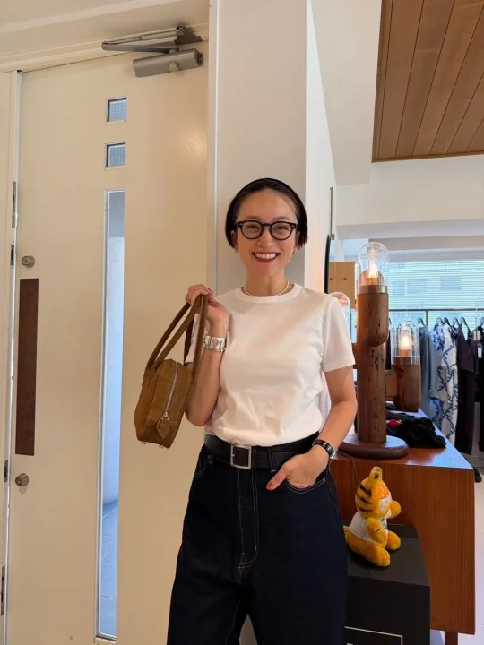 白いTシャツと濃い色のデニムパンツを着用し、眼鏡とパールネックレスをつけた女性