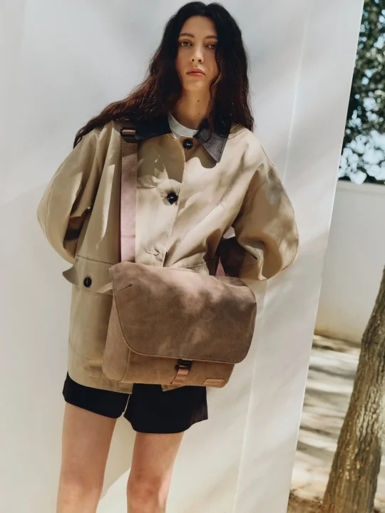 ベージュのジャケットと黒のショートパンツを着用し、ブラウンのメッセンジャーバッグを肩にかけた女性がカメラを見つめて立っています。