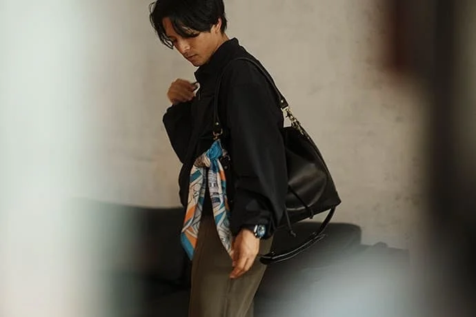 黒いジャケットとオリーブ色のパンツを着用した男性が、ショルダーバッグとスカーフをアクセントにしたカジュアルな着こなし